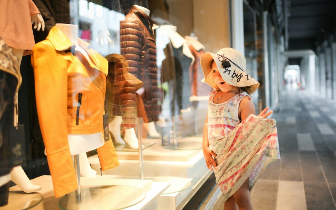 Funny child girl in dress and straw hat looks glass shop, fashion